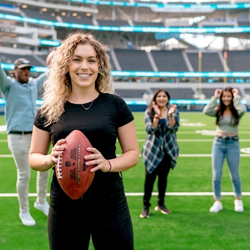 A person holding a football on a field, with three individuals cheering in the background inside a stadium.