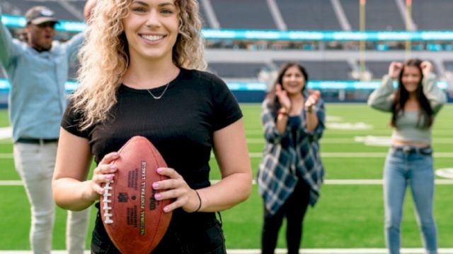 A person holding a football on a field, with three individuals cheering in the background inside a stadium.