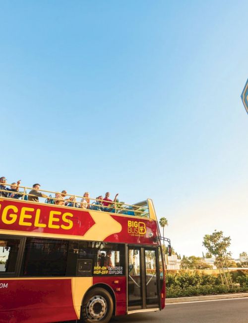 A red double-decker tour bus labeled "LOS ANGELES" near a Beverly Hills sign under a clear blue sky.