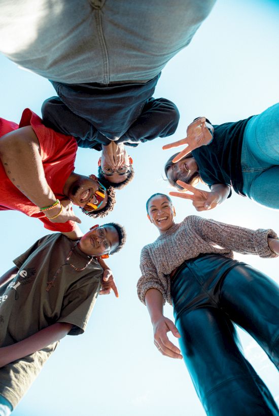 A group of five people stand in a circle, looking down at the camera. They are smiling and making peace signs.