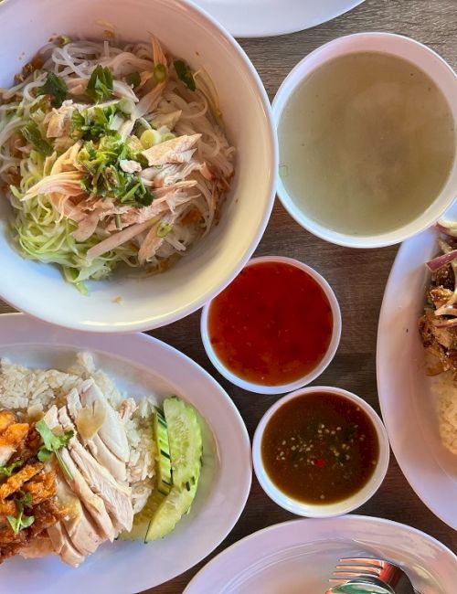The image shows a variety of Asian dishes, including noodles, soup, chicken with rice, and dipping sauces on a wooden table.