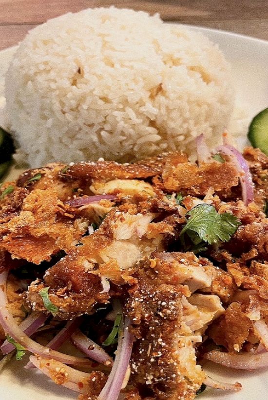 The image shows a plate of crispy chicken with sliced red onions, herbs, rice, and cucumber slices.